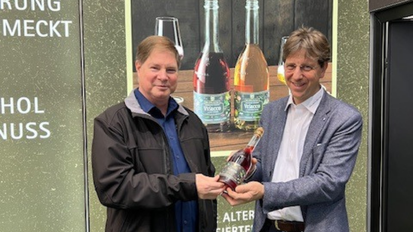 Two men holding a non-alcoholic beverage bottle standing in front of a banner with bottles and wine glasses.