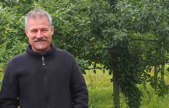 An older gentleman standing in an orchard where the trees are filled with leaves.